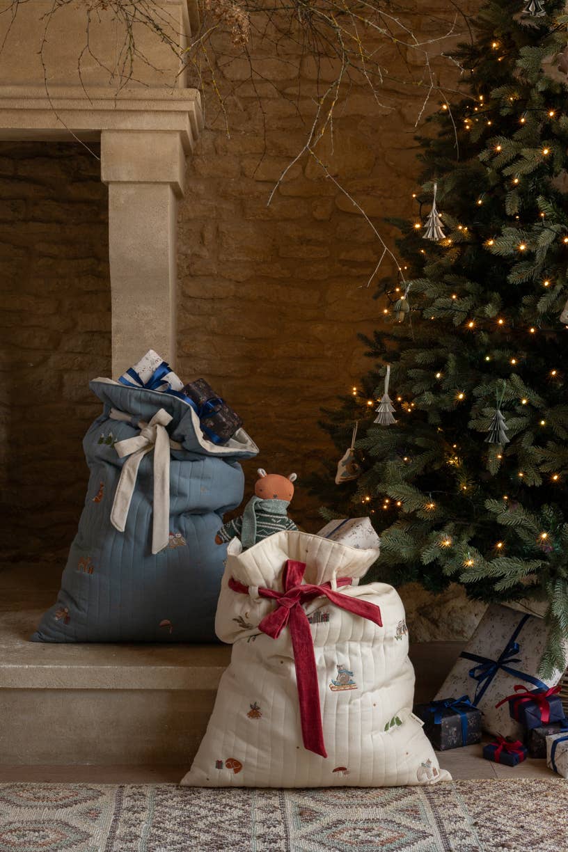 Two fabric gift sacks, one blue and one white with a red bow, in front of a decorated Christmas tree.