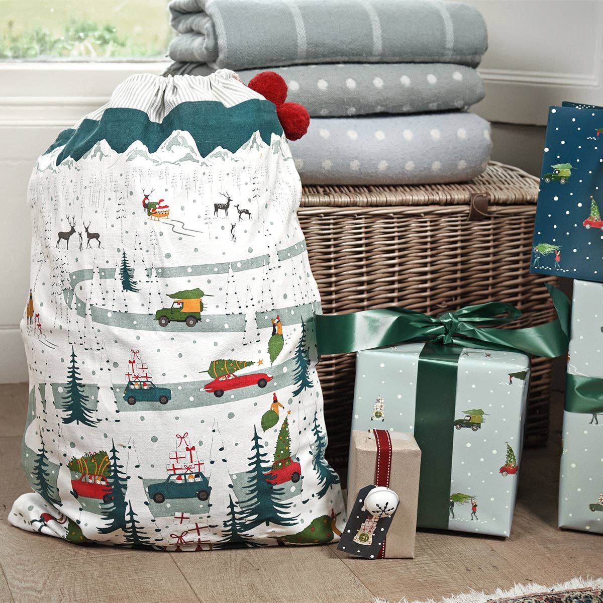 Christmas-themed laundry bag with holiday designs next to wrapped gifts and a wicker basket.