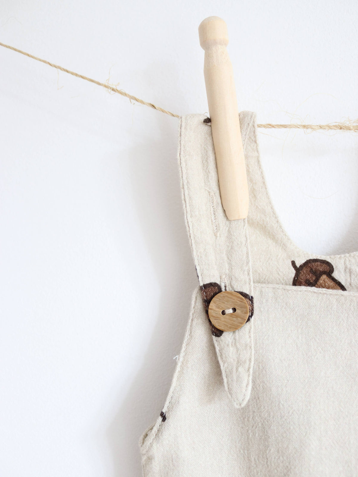 lose-up of beige baby dungarees with a brown acorn print, showing a shoulder strap fastened with a round wooden button.