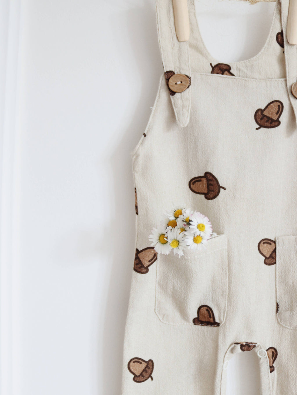 Close-up of beige baby dungarees featuring a brown acorn print, wooden-look shoulder buttons and front pockets decorated with small white and yellow daisies.