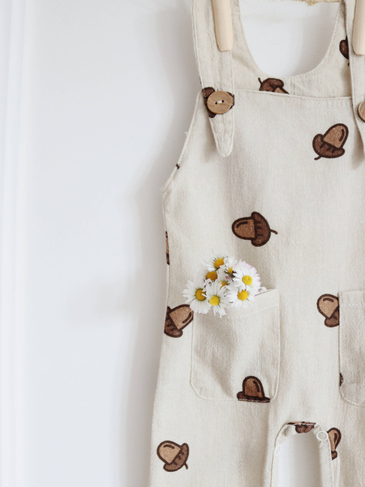 Close-up of beige baby dungarees featuring a brown acorn print, wooden-look shoulder buttons and front pockets decorated with small white and yellow daisies.