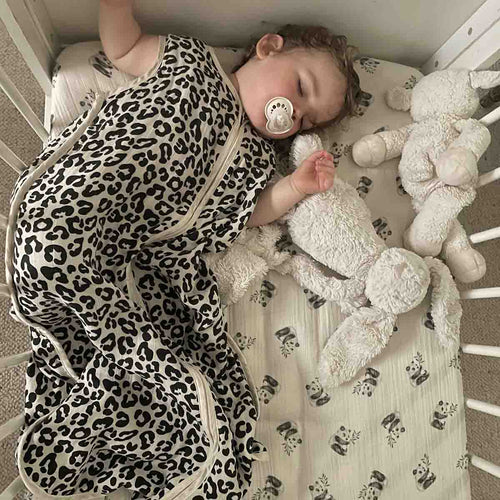 Baby sleeping in the Graou leopard-print Adele sleeping bag in a cot with neutral patterned bedding and soft white plush toys.