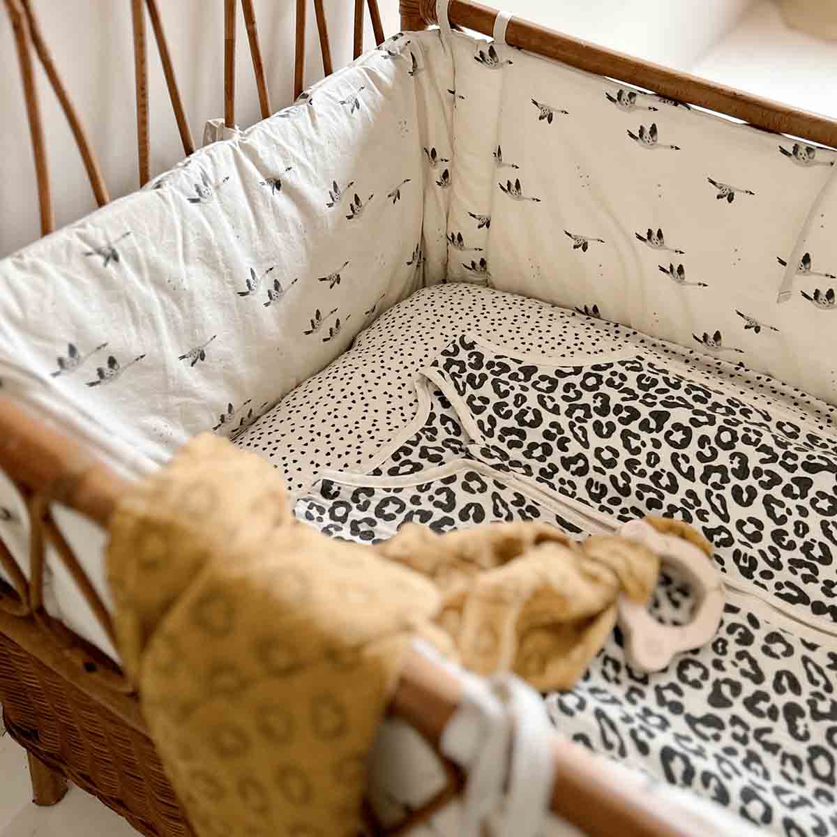 Graou leopard-print baby sleeping bag laid in a wicker bassinet with cream goose-print cot bumpers and neutral nursery bedding.