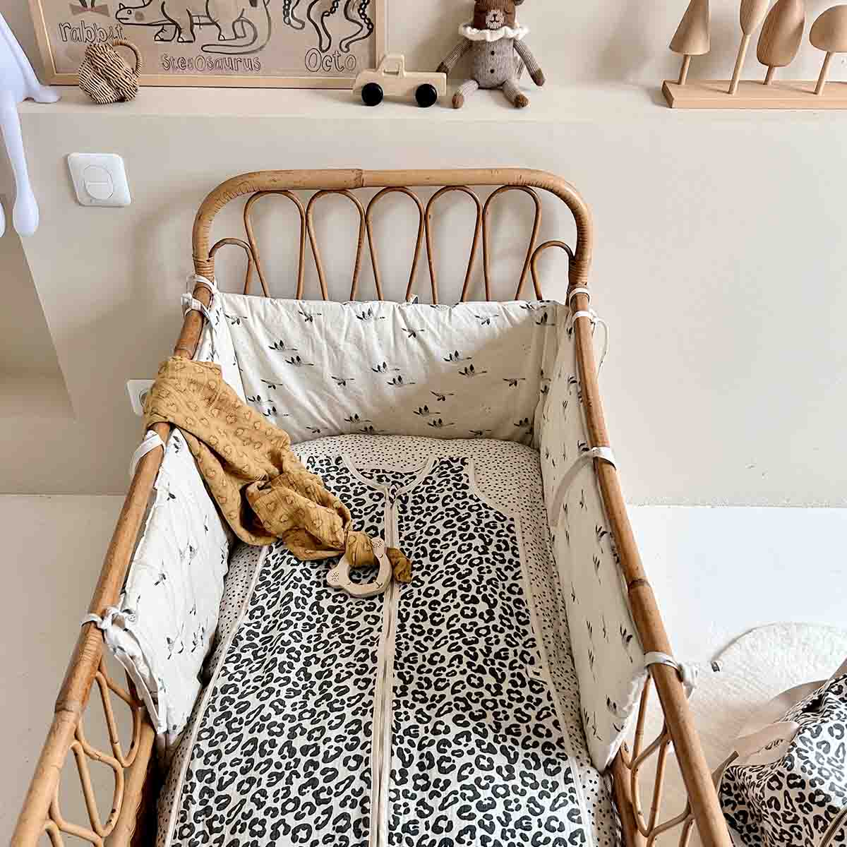 Adele Graou leopard-print baby sleeping bag displayed in a wicker cot with matching patterned bedding and a mustard muslin blanket.