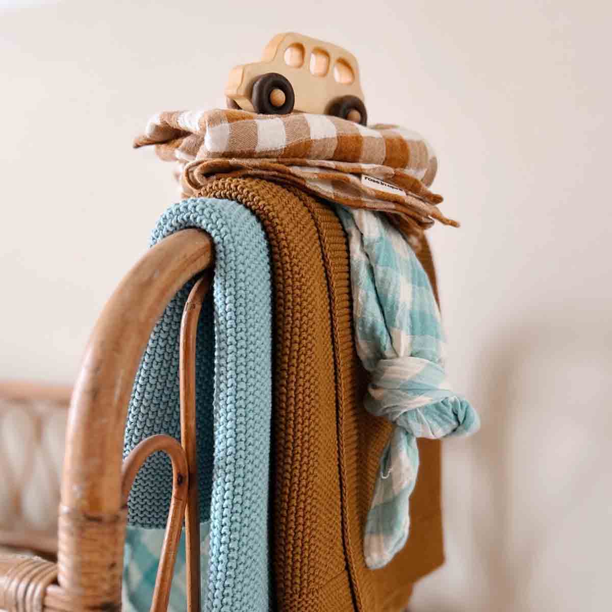 Cumin-brown knitted baby blanket draped over a rattan chair alongside blue and patterned blankets and a wooden toy car.