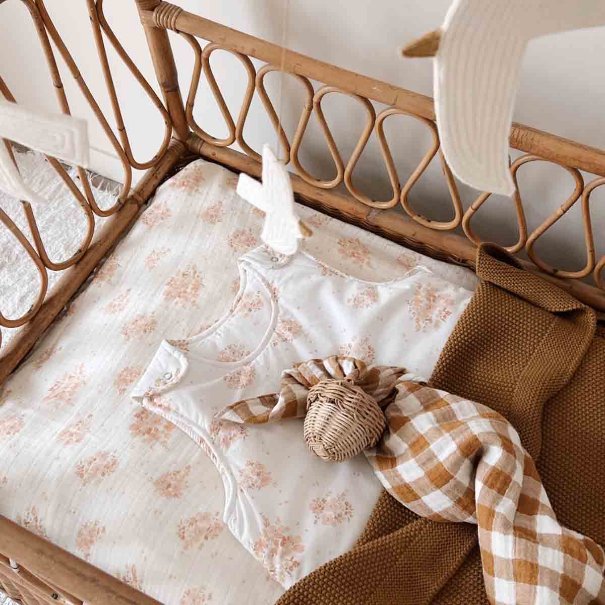 Cumin-brown knitted baby blanket arranged in a rattan cot with neutral floral bedding, a checked muslin cloth and a soft baby vest.