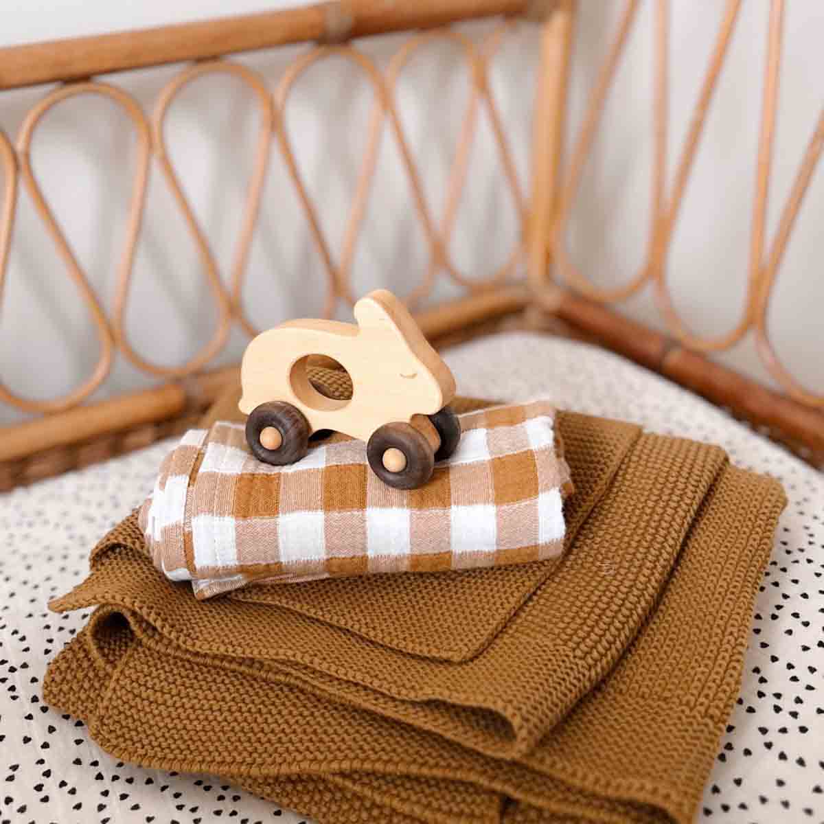 Folded cumin-brown knitted baby blanket styled in a rattan cot with a wooden toy car and a brown-and-white checked muslin cloth.