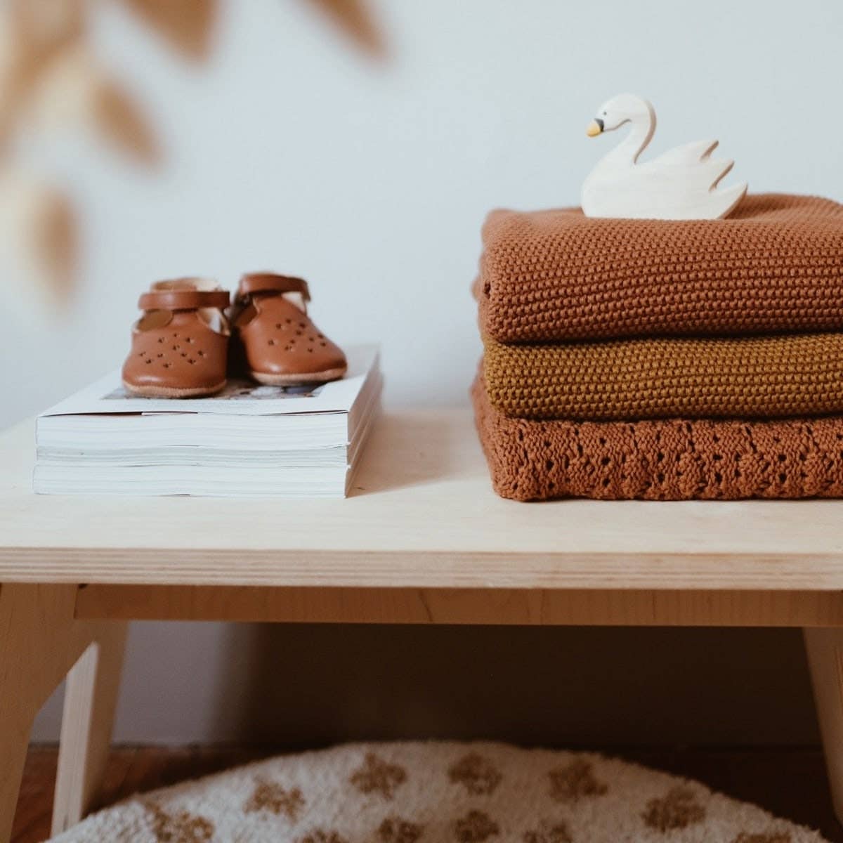 Stack of warm brown knitted baby blankets with a white swan ornament and tiny brown baby shoes on a wooden table.