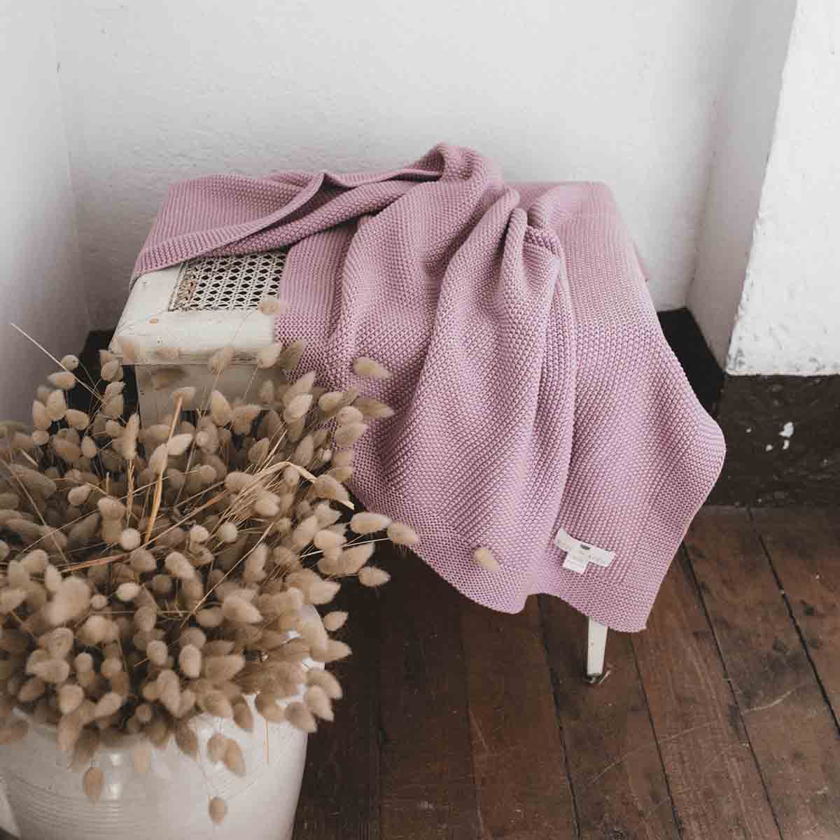 Stack of knitted cotton blankets in mustard yellow, navy, lilac and pale blue displayed on a white stool.