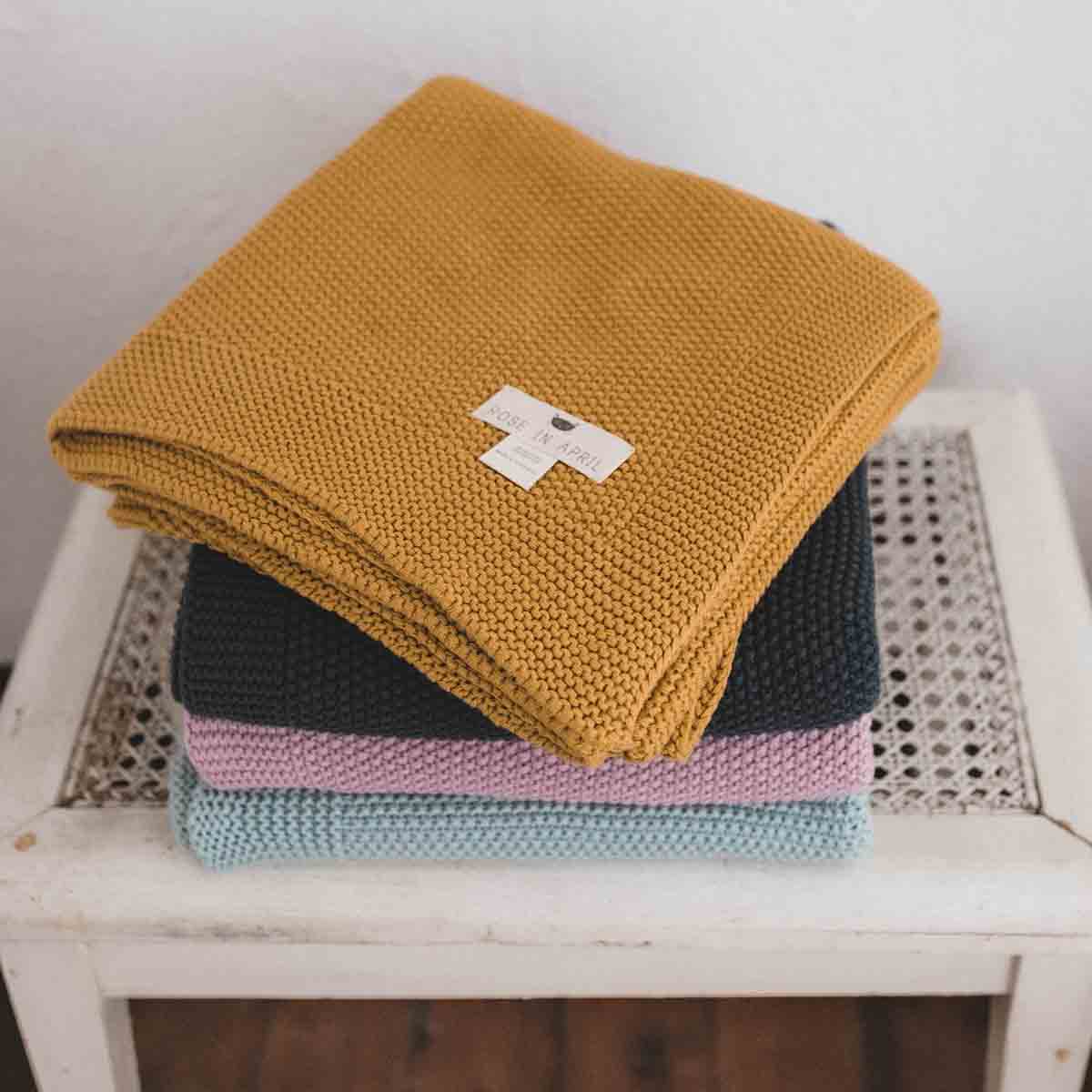 Stack of knitted cotton blankets in mustard yellow, navy, lilac and pale blue displayed on a white stool.