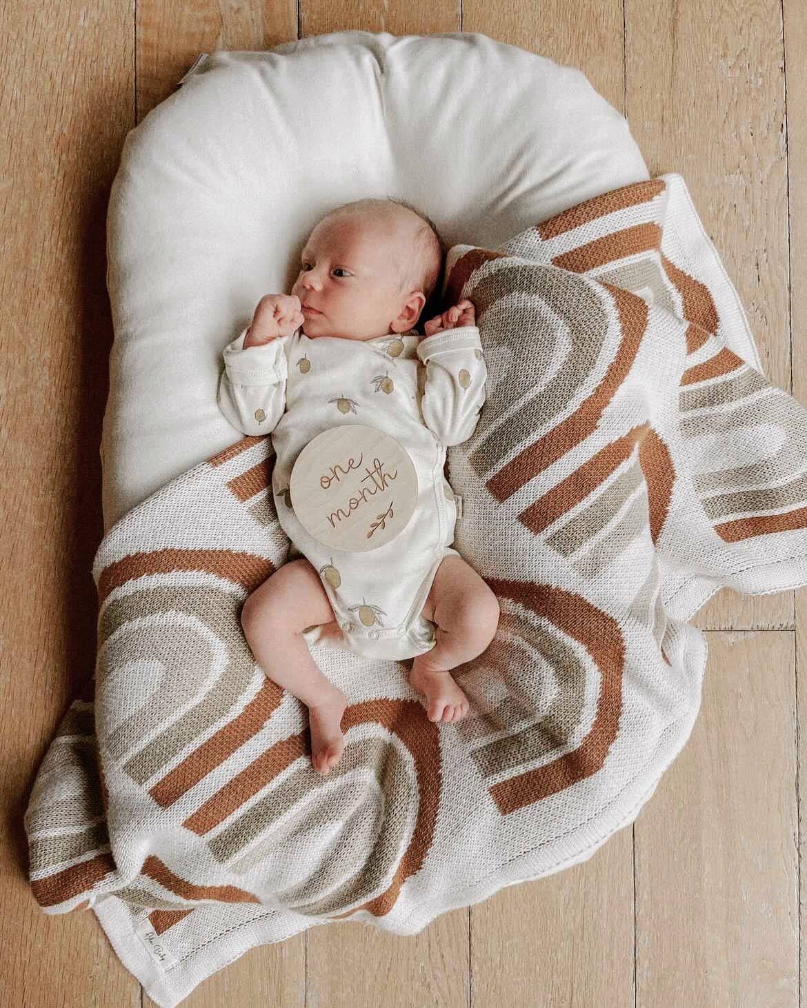 Baby lying on a cream knitted blanket with a rust and beige rainbow pattern, wearing a cream sleepsuit and holding a round one-month milestone disc.