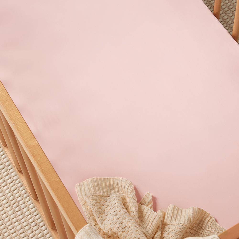 Close-up of a smooth baby pink fitted cot sheet on a wooden cot, styled with a cream knitted blanket.