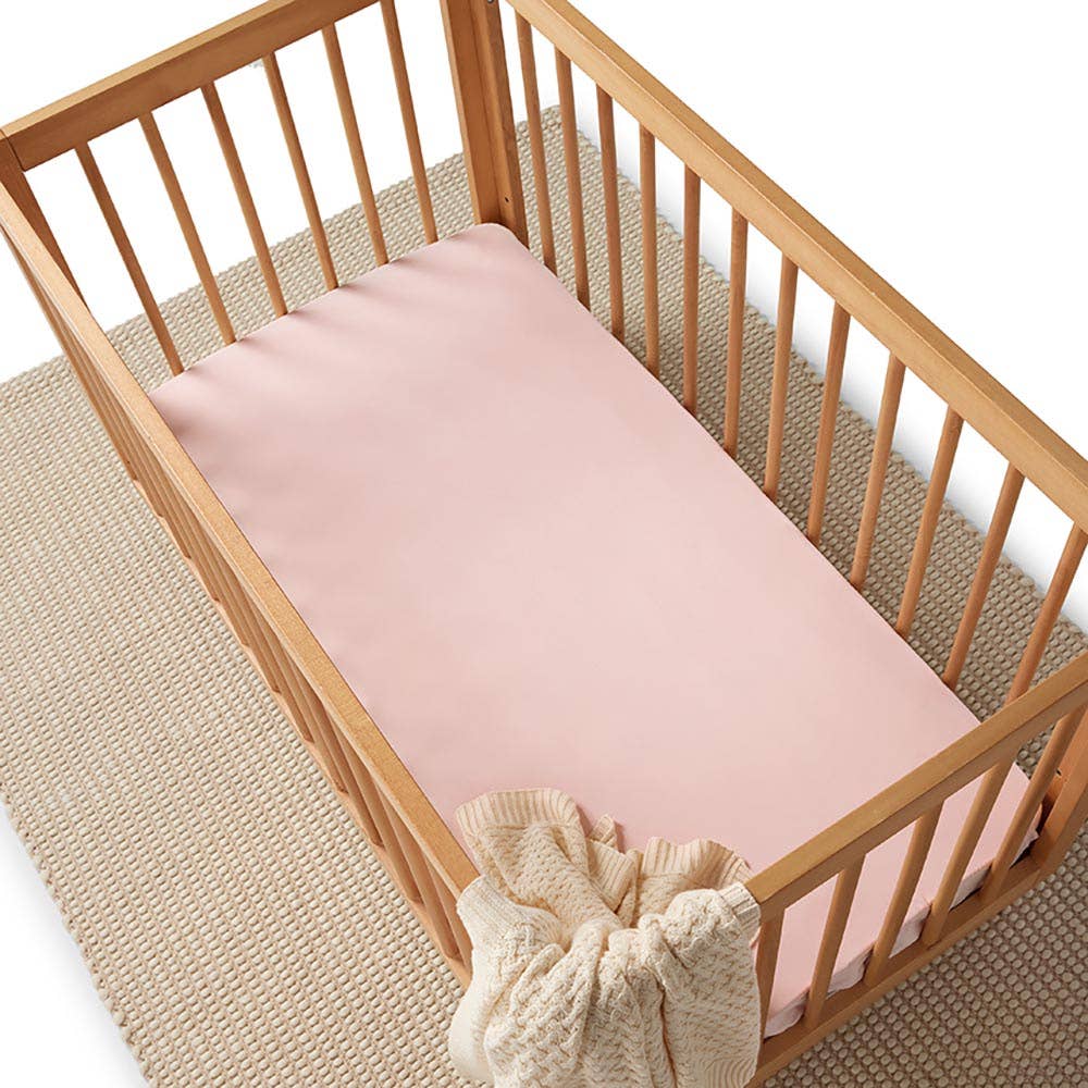 Wooden cot with a baby pink fitted cot sheet and a cream knitted blanket draped over the right-hand rail, displayed on a neutral woven rug.