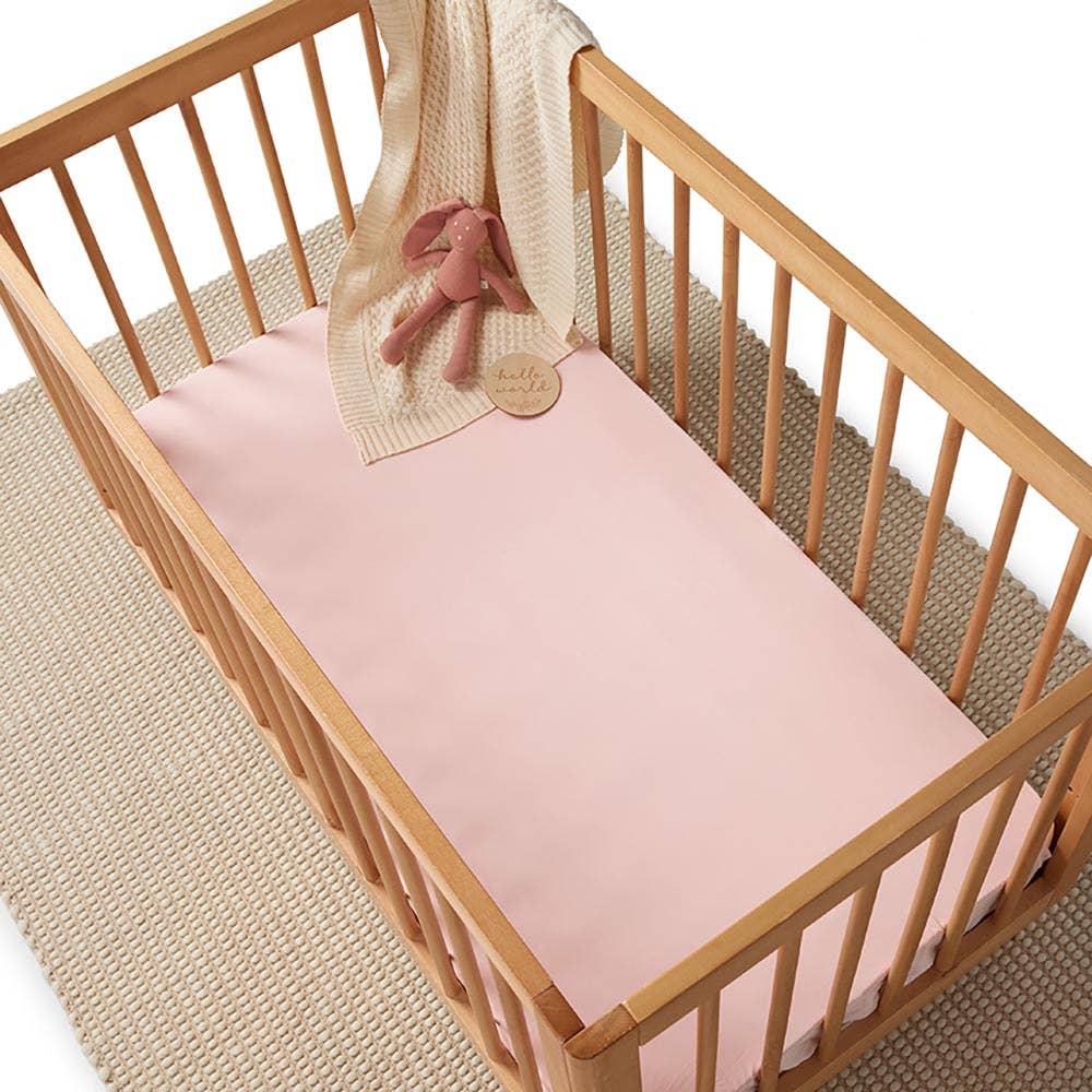 Baby pink fitted cot sheet covering a wooden cot mattress, styled with a cream knitted blanket and pink bunny soft toy.