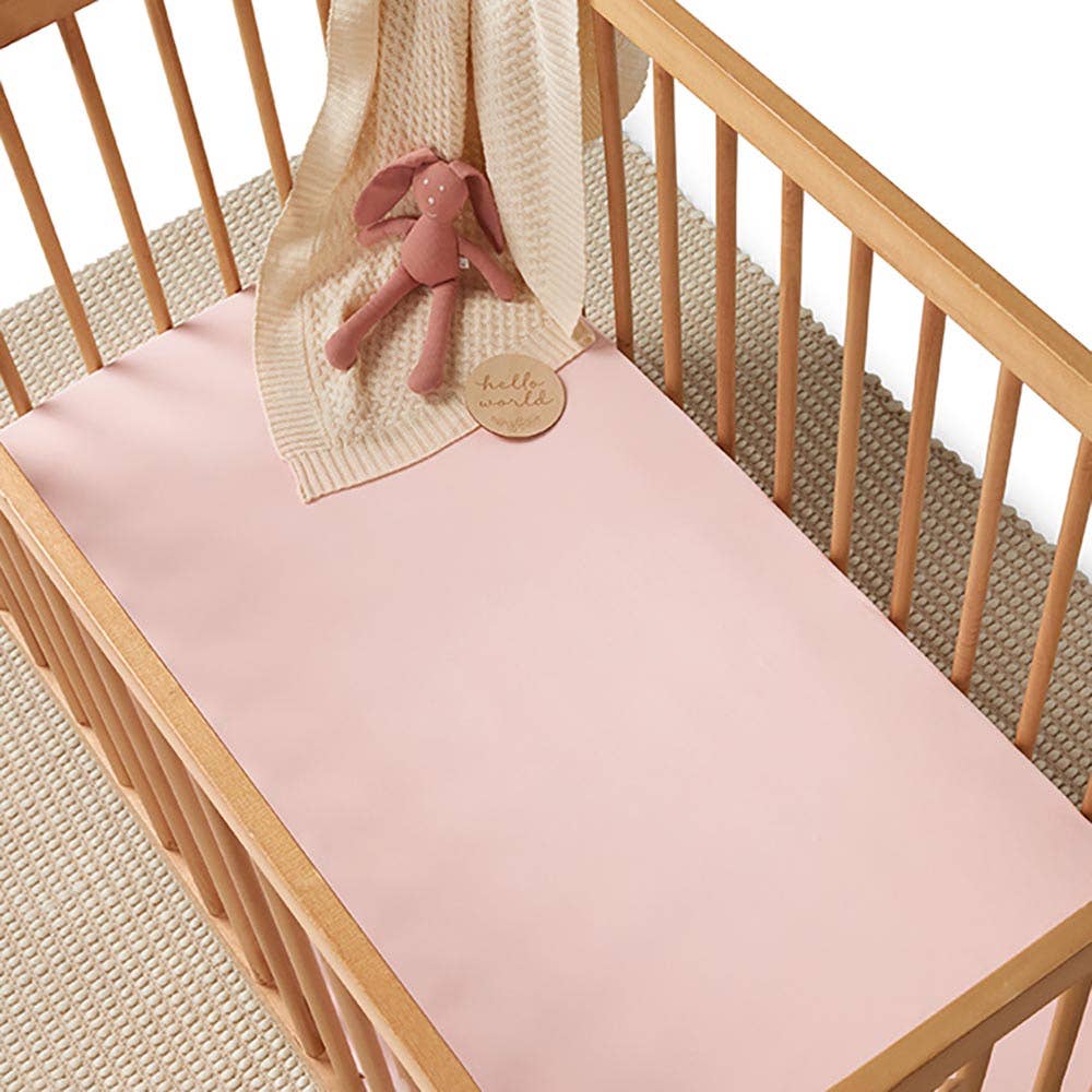 Baby pink fitted cot sheet covering a wooden cot mattress, styled with a knitted cream blanket and a pink bunny soft toy.
