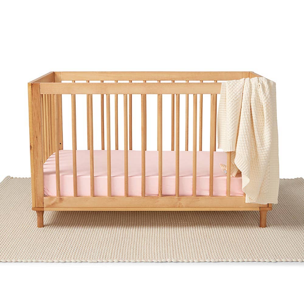 Wooden cot with a baby pink fitted cot sheet and a cream knitted blanket draped over the side, shown against a neutral nursery background.