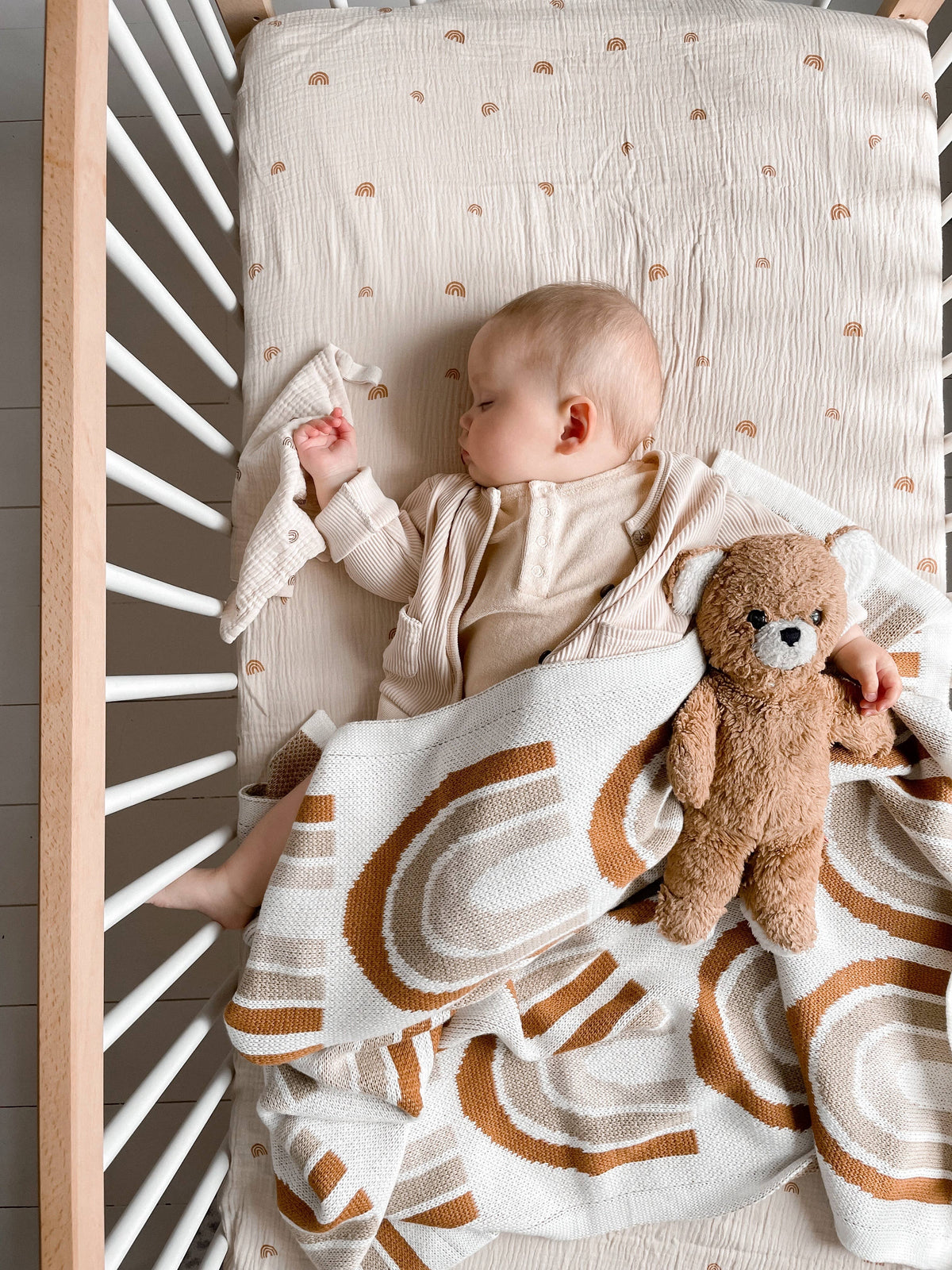 Baby lying in a white cot wrapped in a cream knitted blanket with a rust and beige rainbow pattern, beside a brown teddy bear.