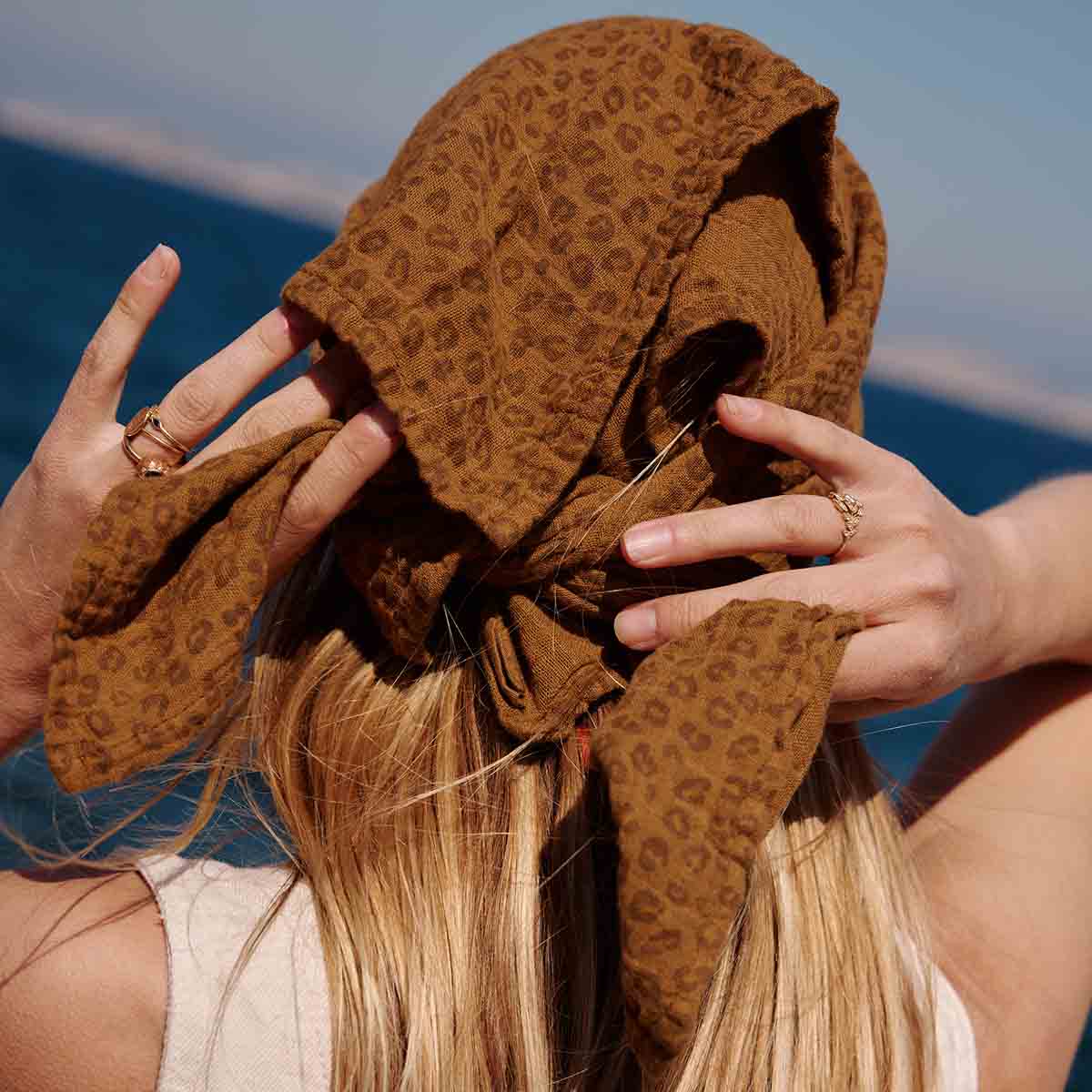 Close-up of a woman tying the brown leopard-print muslin scarf around her head.