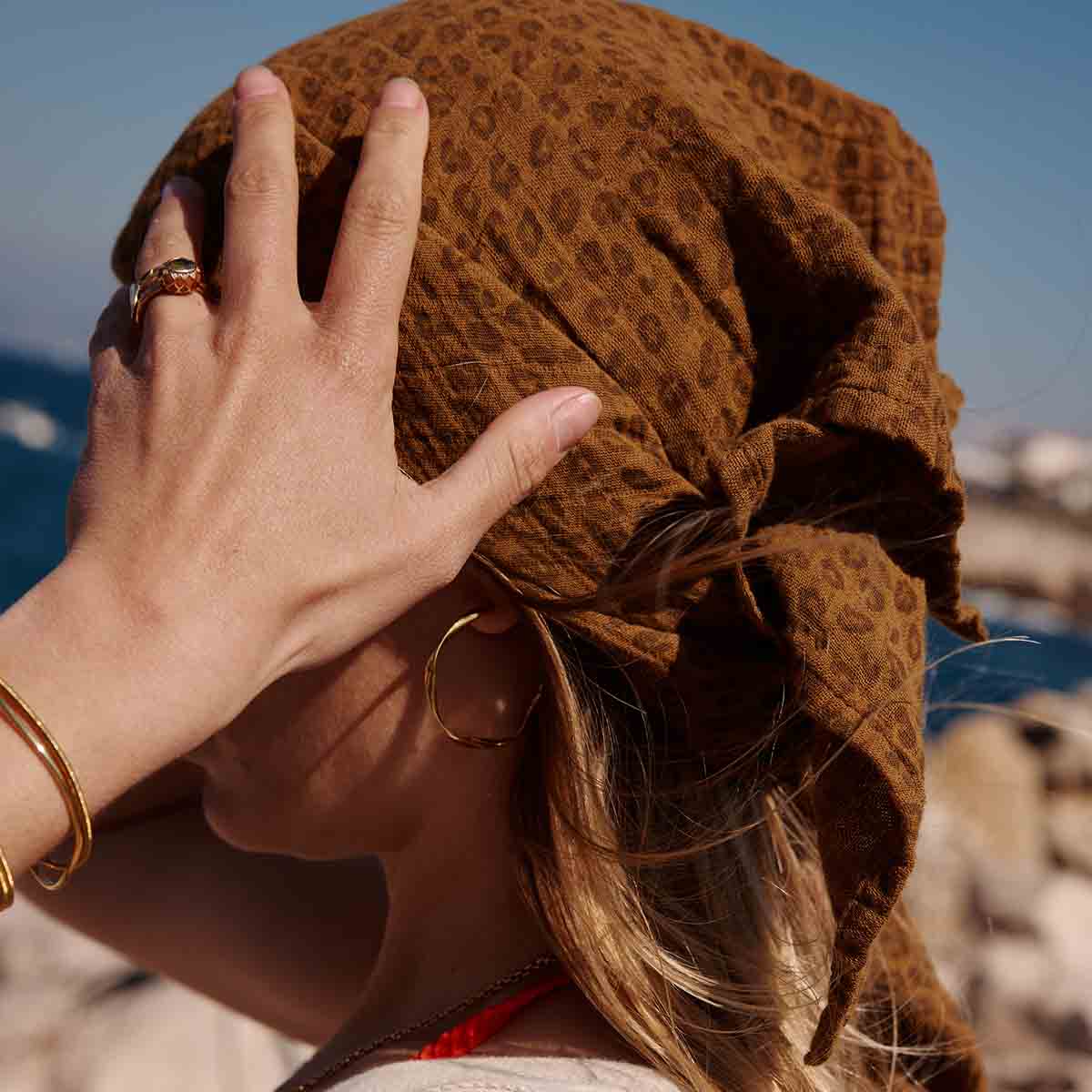 Woman wearing the brown Mini Leopard Cumin muslin scarf over her head outdoors by the sea.