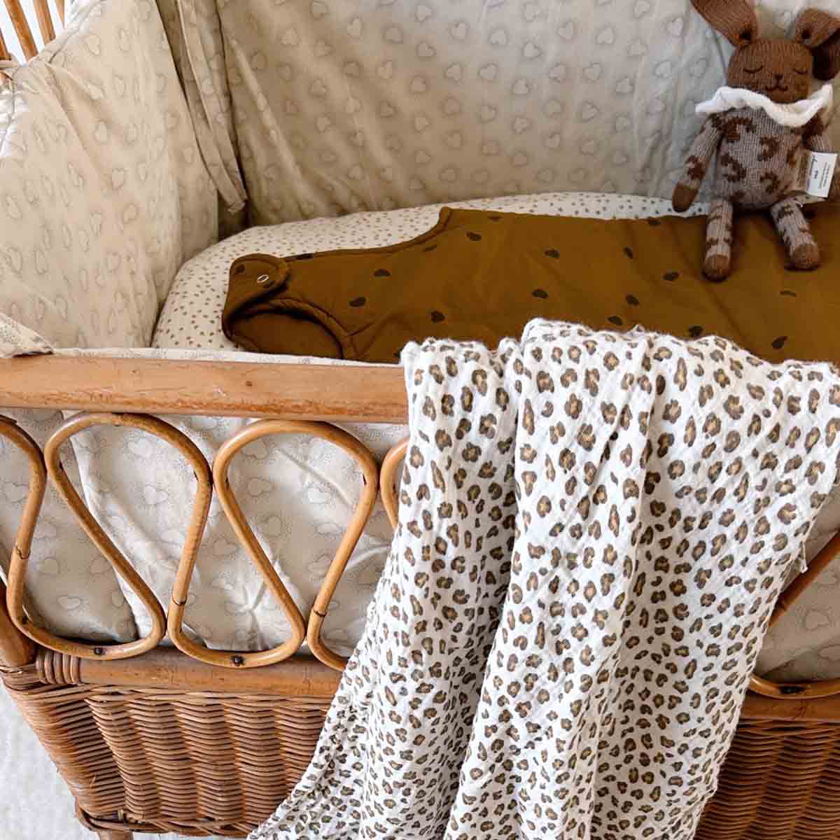 Bianca Mini Leopard Greige muslin swaddle draped over a wicker bassinet, styled with neutral nursery bedding and a brown rabbit soft toy.