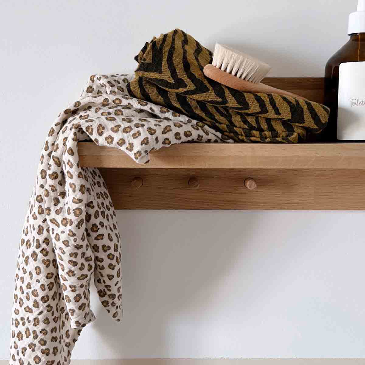 Bianca Mini Leopard Greige muslin swaddle draped over a wooden shelf beside a wooden baby brush and amber bottle.