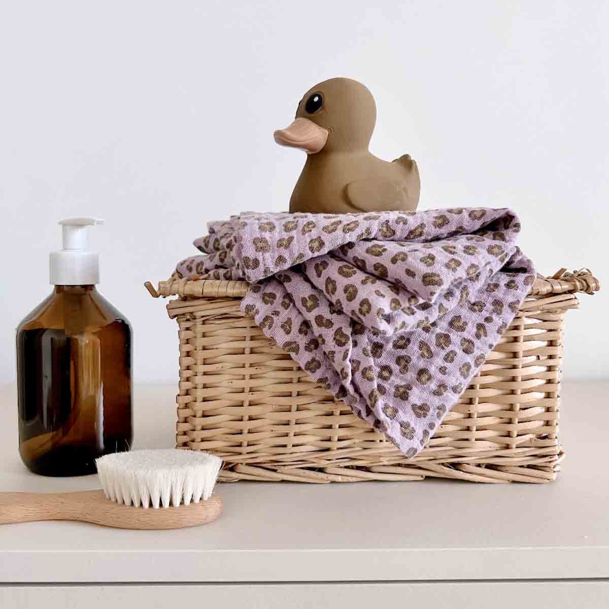 Lilac leopard-print muslin swaddle draped over a wicker basket beside a rubber duck and a baby grooming brush.