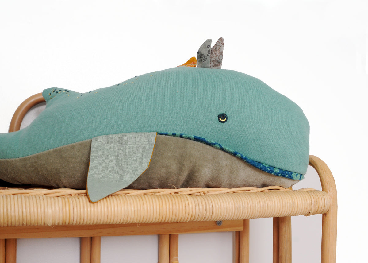 Close-up of a blue whale cushion with embroidered details displayed on a rattan nursery shelf