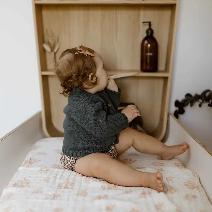 Baby sitting on a cream changing mat cover with a peach bouquet print in a wooden changing area.