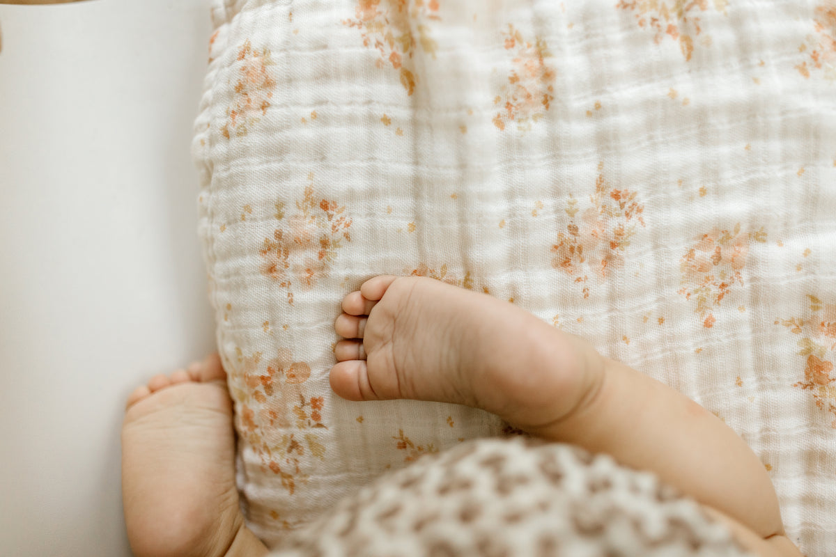 Close-up of a baby’s feet resting on a cream muslin changing mat cover with delicate peach floral prints.