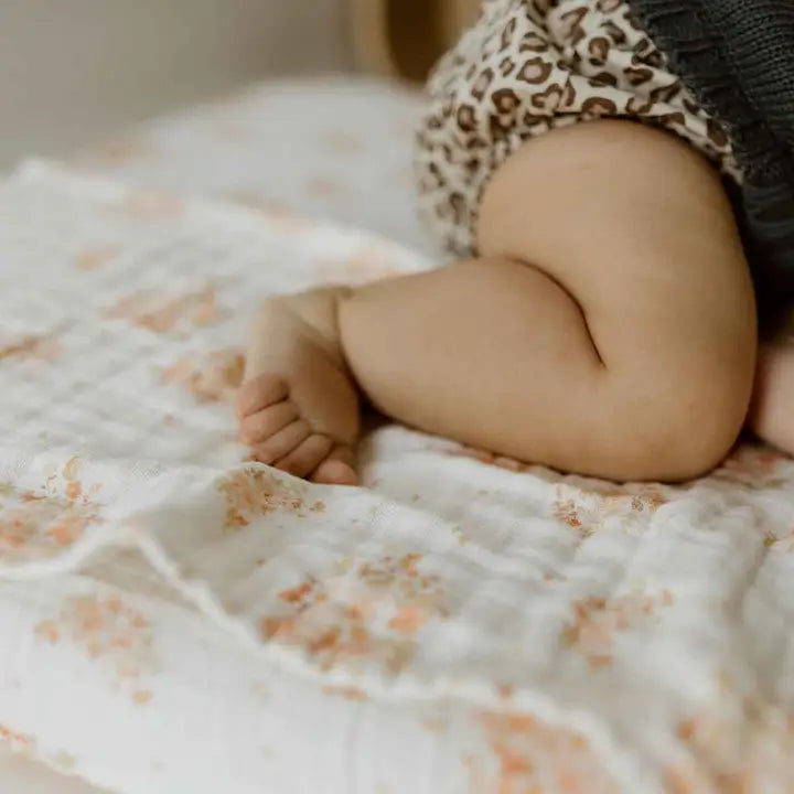 Close-up of a baby’s legs on a cream muslin changing mat cover featuring a peach and pink floral bouquet pattern.