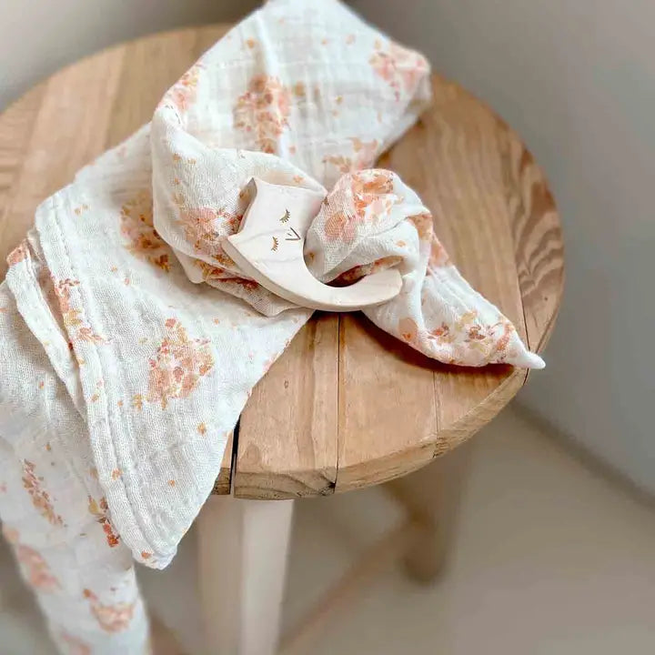 Cream muslin blanket with delicate peach and pink floral bouquet prints draped over a wooden table beside a wooden baby teether.