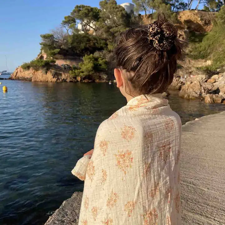 Woman wearing a cream muslin blanket with a peach floral print draped around her shoulders beside the sea.