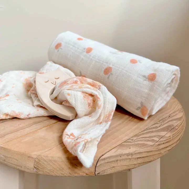 Cream cotton muslin blanket with peach floral and dot prints, rolled beside a wooden baby teether on a round wooden table.