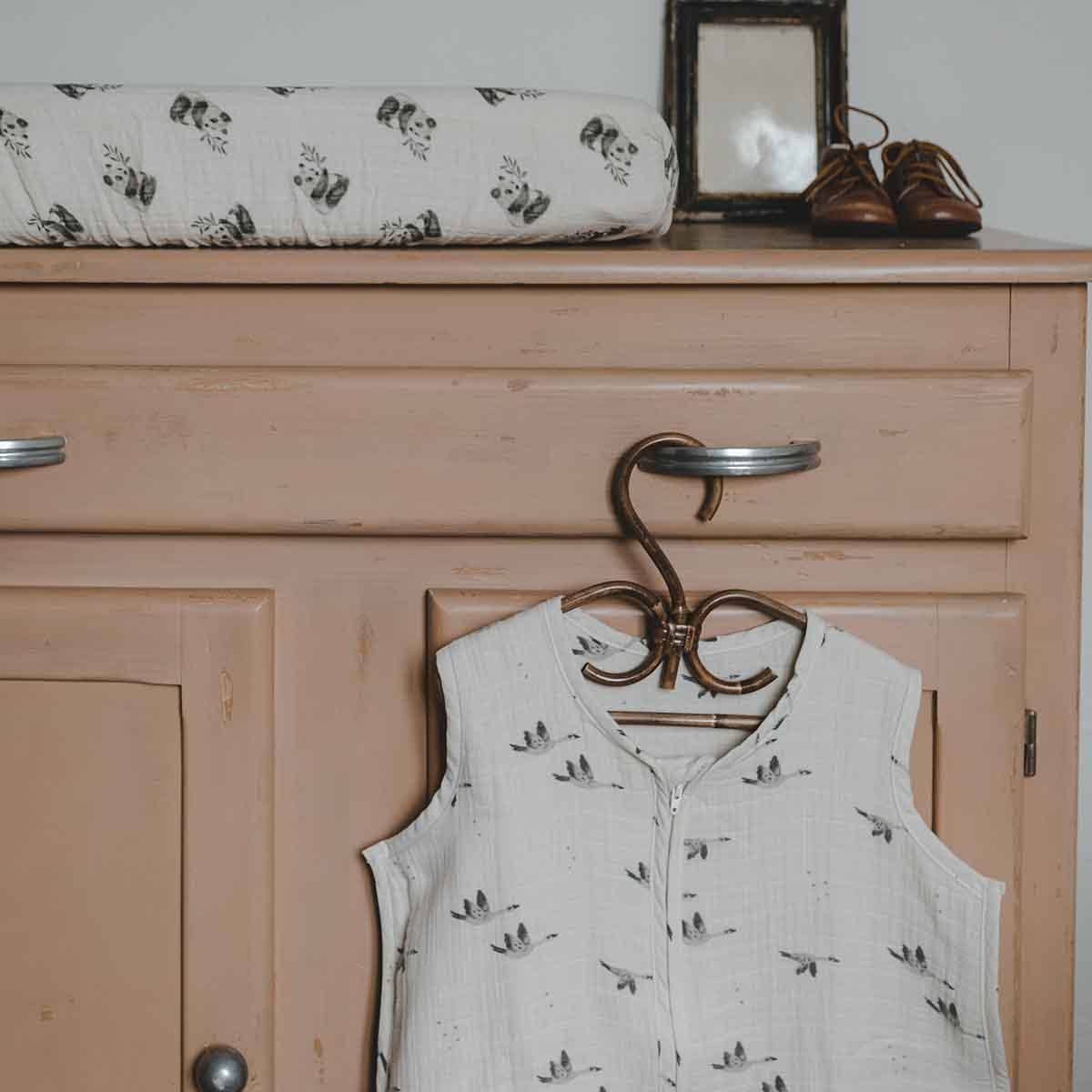 Charly Goose lightweight summer baby sleeping bag in cream with a grey flying goose print, hanging from a wooden hanger on a pink cabinet.