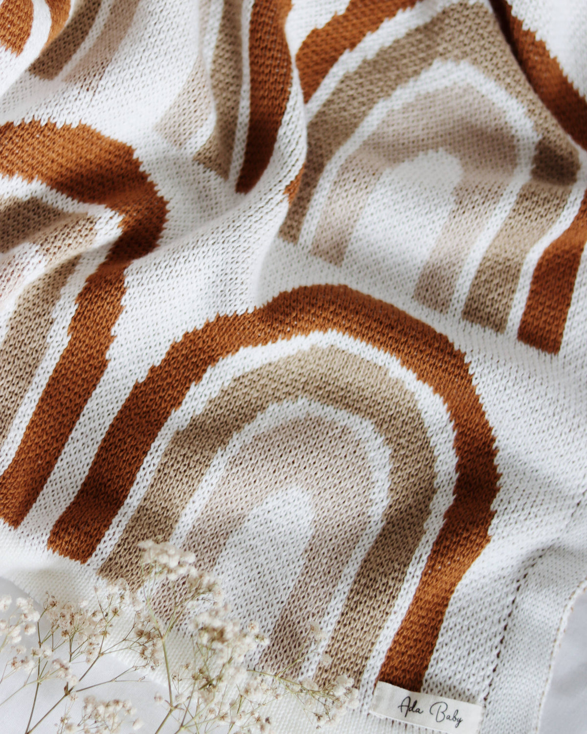 Close-up of a cream knitted blanket featuring a rust and beige rainbow pattern, showing the textured knit and woven fabric label.