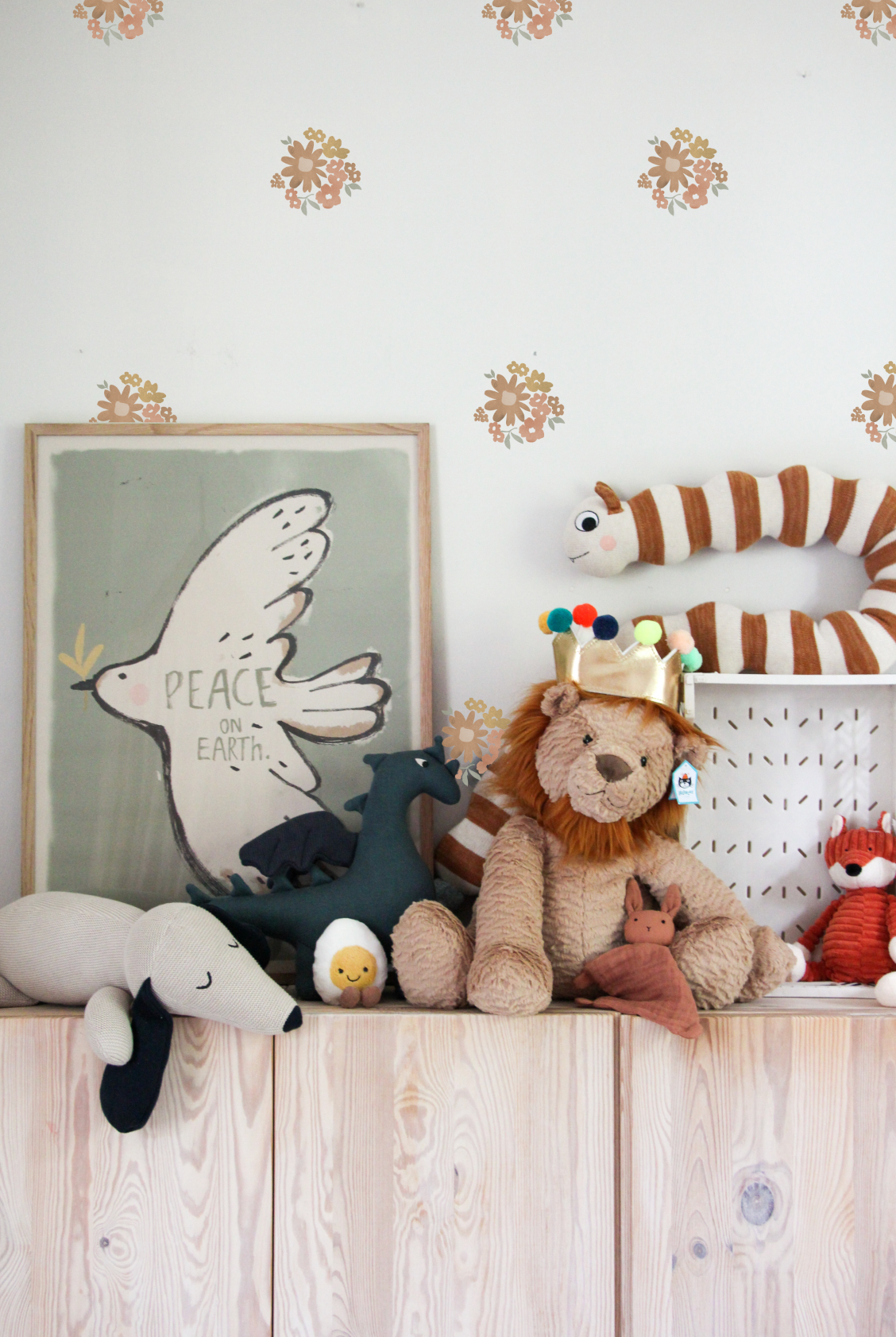 Ditsy floral wall stickers in muted mustard, terracotta and sage green displayed above a nursery shelf with plush animal toys, a framed “Peace on Earth” bird print and a striped tiger soft toy.