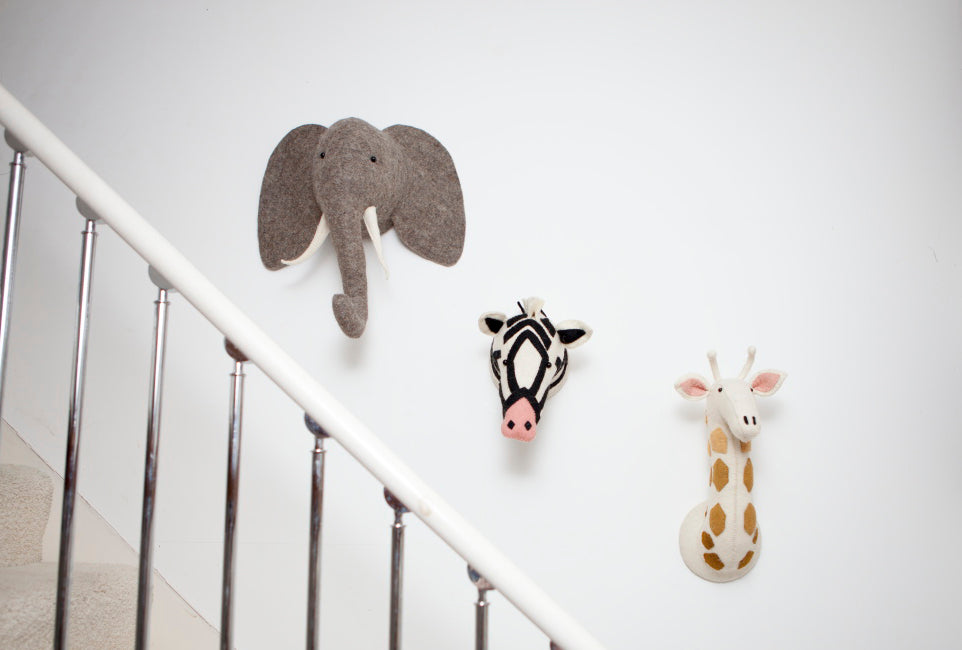 Fiona Walker grey elephant head wall décor displayed on a white wall beside zebra and giraffe animal heads along a staircase, creating a safari-themed interior.