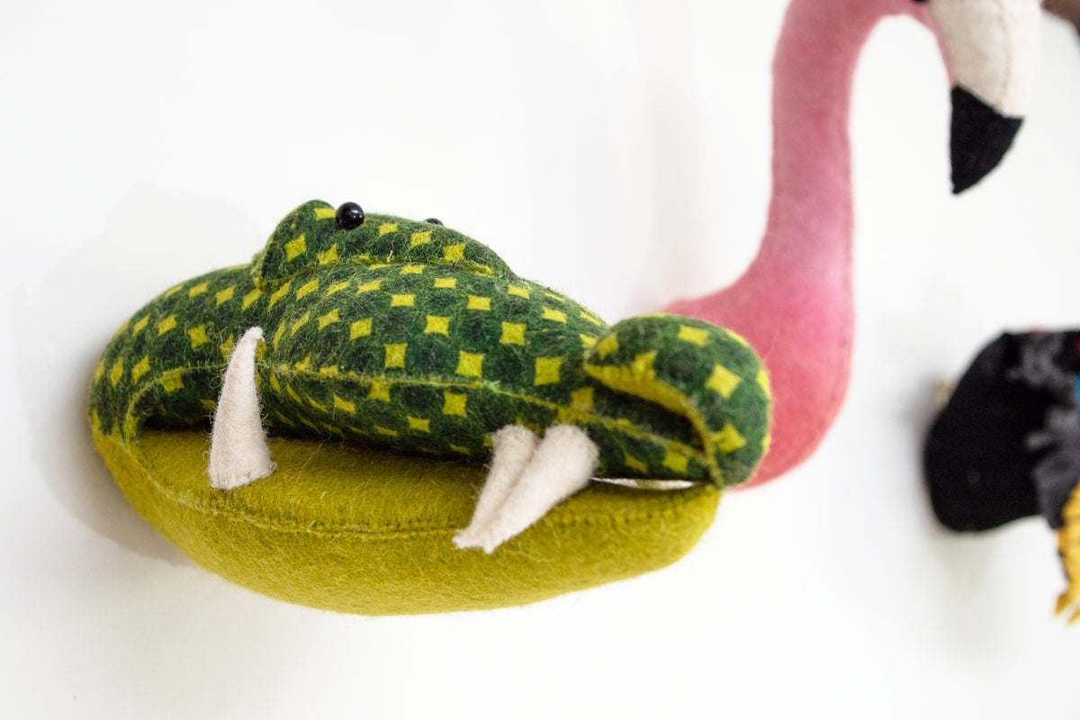 Close-up of the Fiona Walker mini alligator head wall décor, featuring a green and lime checkered wool surface, black eyes and cream-coloured teeth, displayed beside a pink flamingo wall decoration.