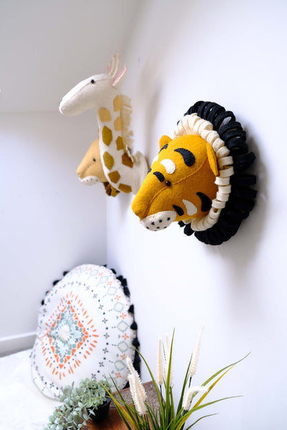 Fiona Walker Roary the Tiger felt wall head displayed beside a giraffe and lion wall head in a safari-themed nursery with a patterned floor cushion.