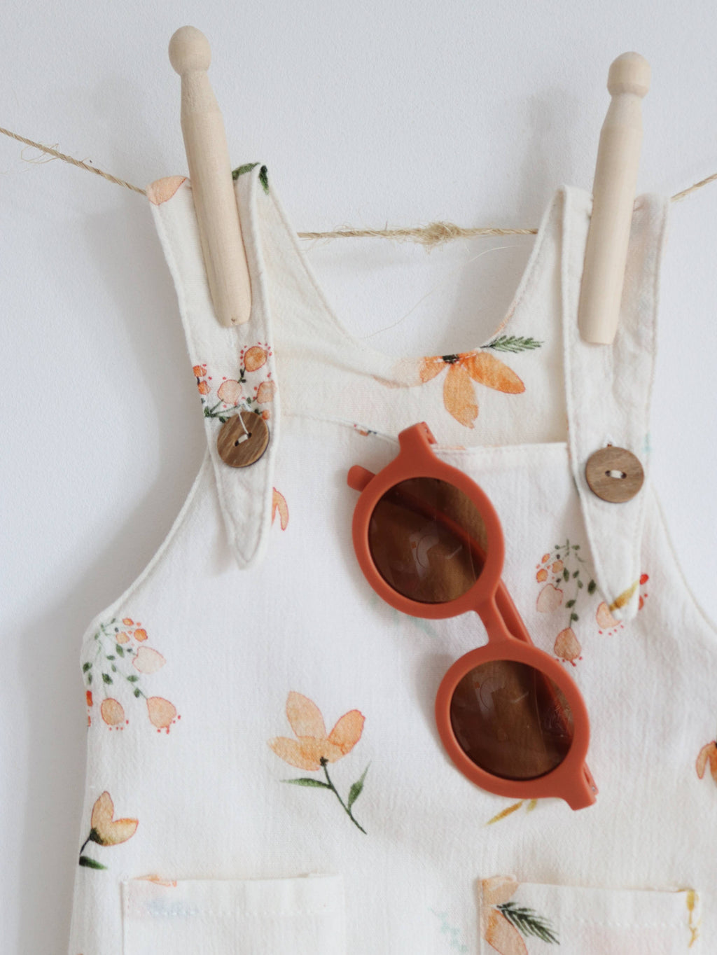 Close-up of cream baby dungarees with a delicate peach and orange floral print, wooden-look shoulder buttons and front pockets, styled with rust-coloured sunglasses.