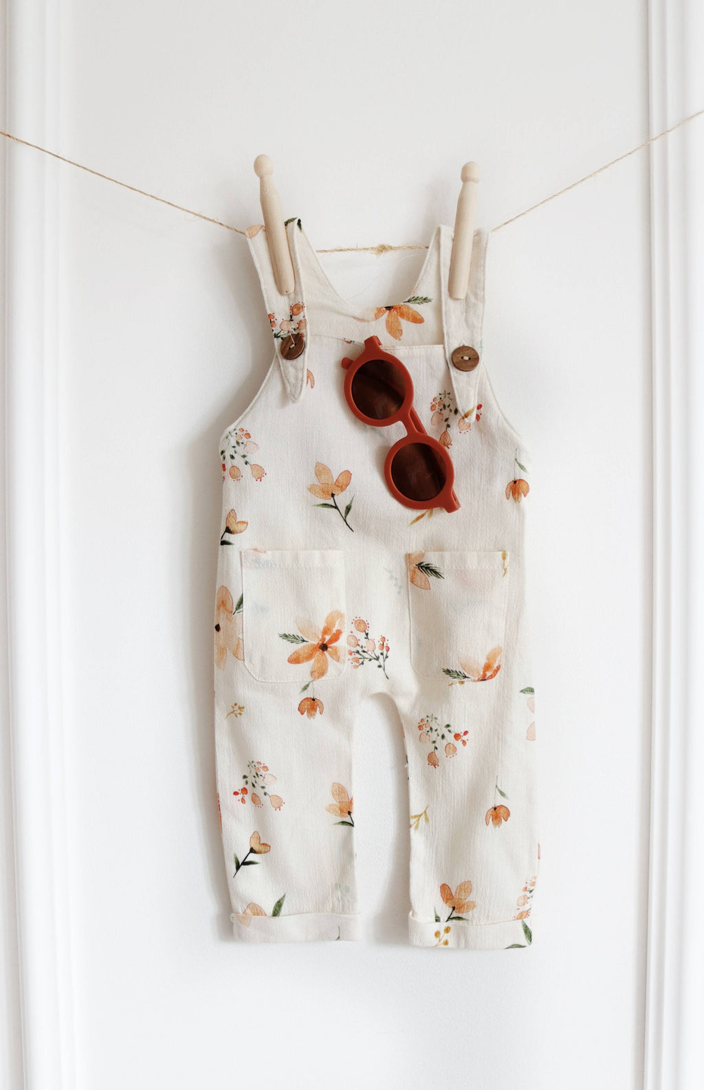 Cream baby dungarees featuring a peach and orange floral print, wooden-look shoulder buttons and two front pockets, styled with rust-coloured sunglasses.