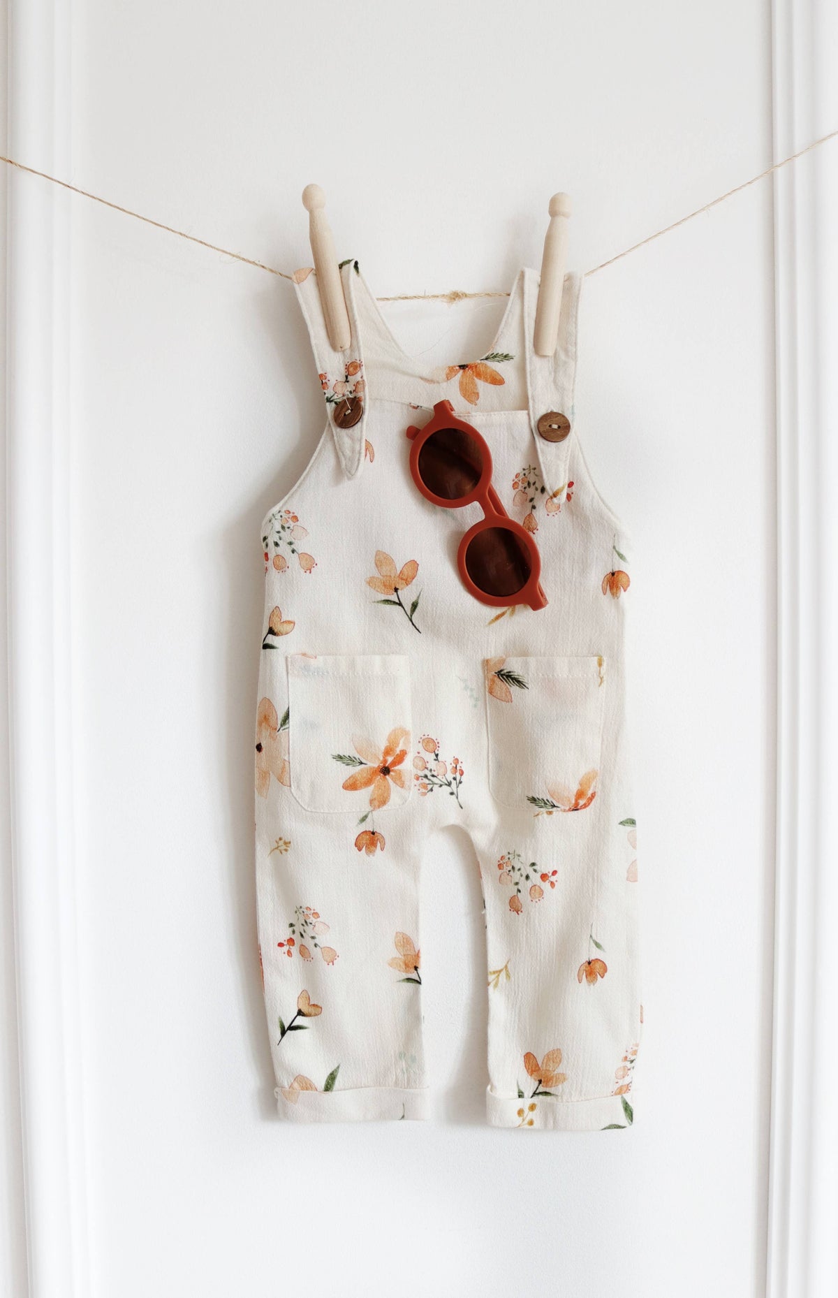 Cream baby dungarees featuring a peach and orange floral print, wooden-look shoulder buttons and two front pockets, styled with rust-coloured sunglasses.