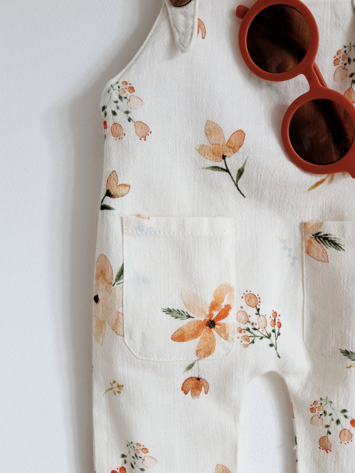 Close-up of cream baby dungarees featuring a peach and orange floral print with green leaves, two front pockets and rust-coloured sunglasses.