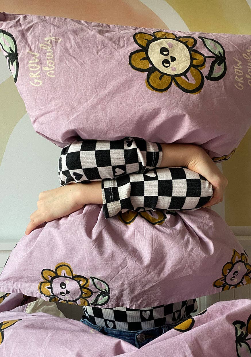 Close-up of a pink floral duvet cover featuring playful yellow flower characters, green leaves and decorative lettering, held by a child wearing black and white checked sleeves.