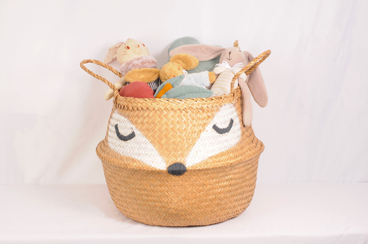 Foxy woven seagrass storage basket with a sleepy fox face and braided handles, filled with colourful plush animals and soft toys against a light background.
