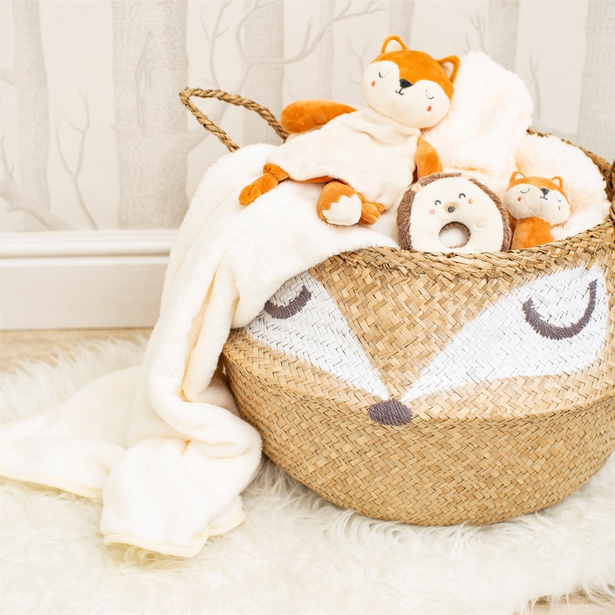 Round woven seagrass Foxy storage basket with a natural brown finish and a sleepy fox face, filled with a cream blanket and woodland-themed soft toys in a nursery.
