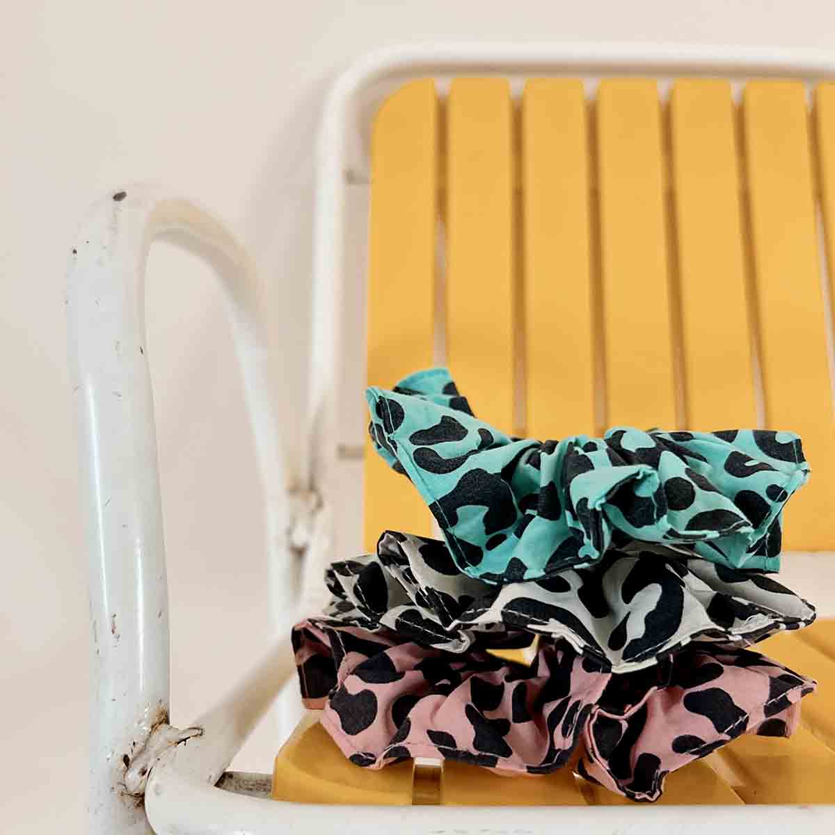 Three leopard-print scrunchies in turquoise, greige and pink stacked on a yellow chair, showcasing the colourful animal-print designs.