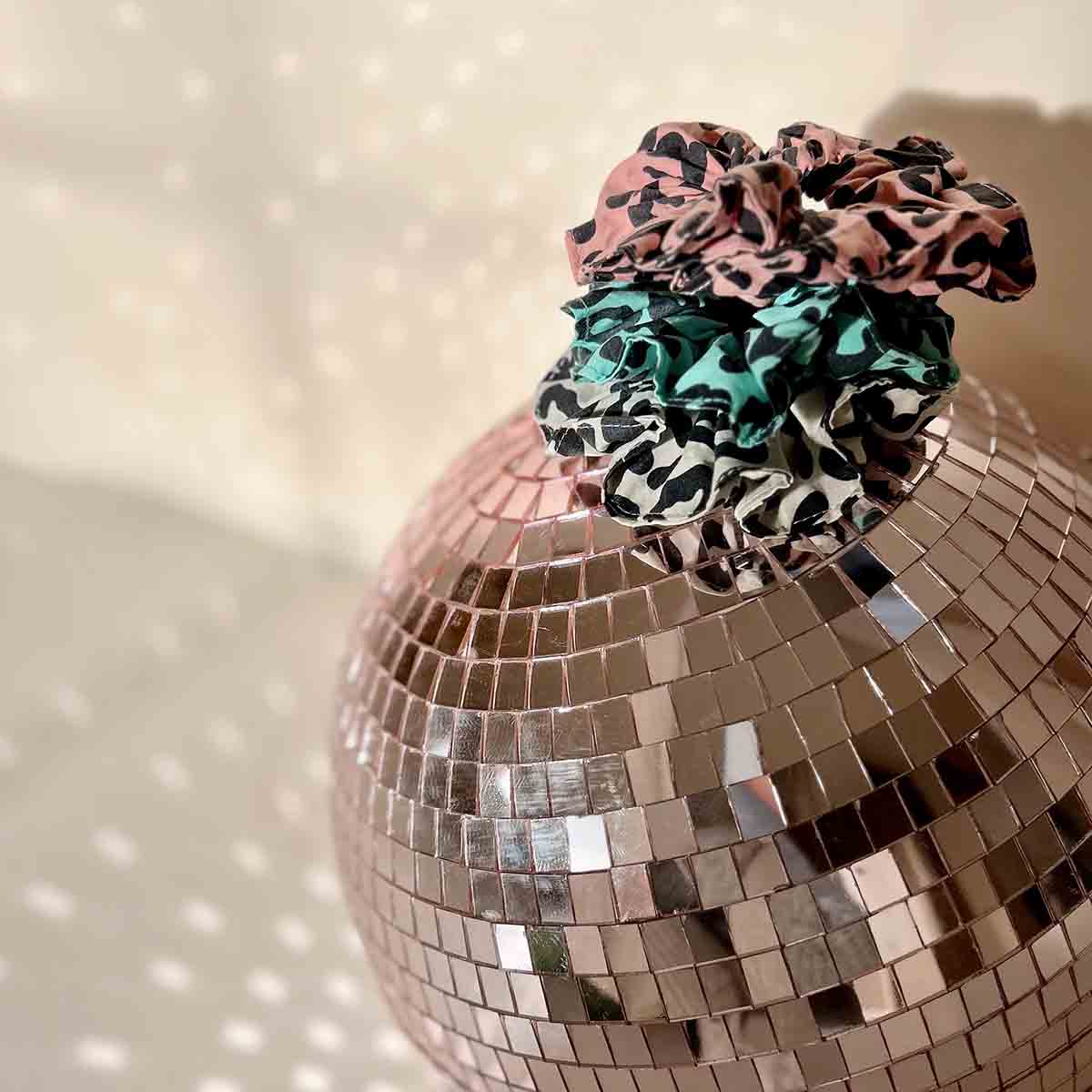 Pink, turquoise and greige leopard-print scrunchies stacked on a mirrored disco ball in warm decorative lighting.