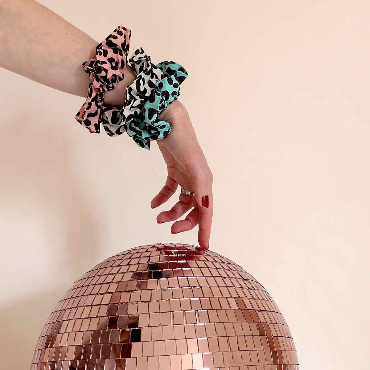 Three leopard-print scrunchies in pink, greige and turquoise worn together on a wrist above a rose-gold mirrored disco ball.