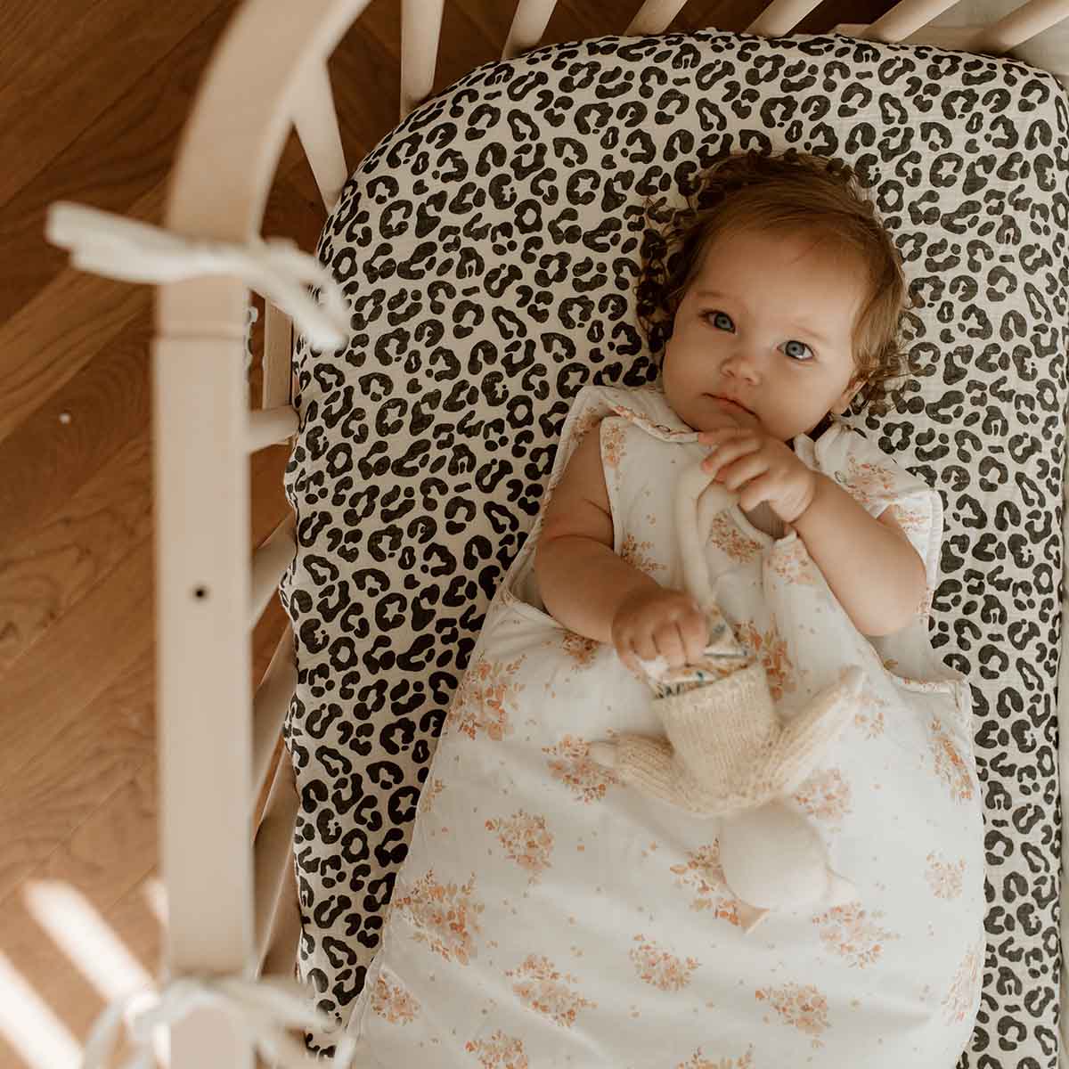 Baby lying in a wooden cot on a cream fitted sheet with a bold black animal print, wearing a cream floral sleeping bag.